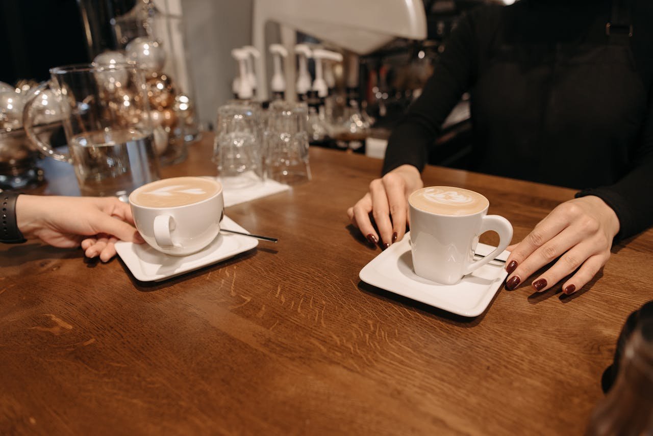 A barista serves two foamy lattes in a cozy coffee shop setting, showcasing latte art.